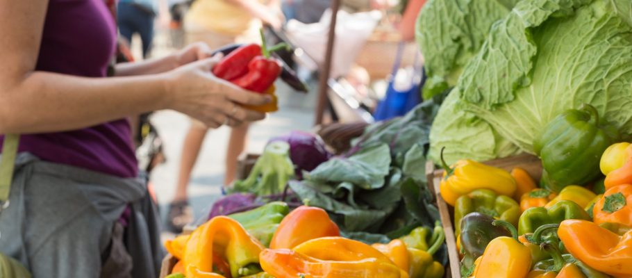 Colorful fresh produce at a farmers market in the foreground with a woman and her purchases in the background.